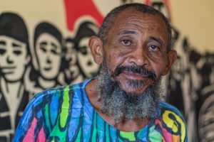 Imagem mostra Nego Bispo, um homem negro, com barba grisalha e cabelo curto e grisalho também. Ele veste uma camiseta colorida.