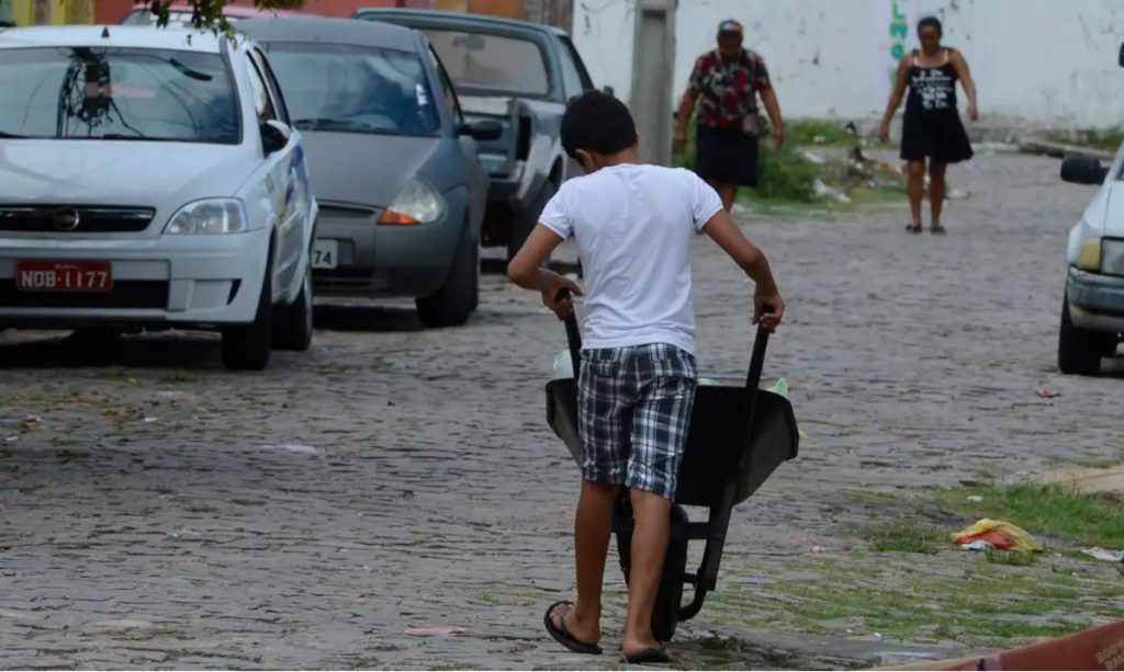 Imagem mostra um menino de costas carregando um carrinho de mão.