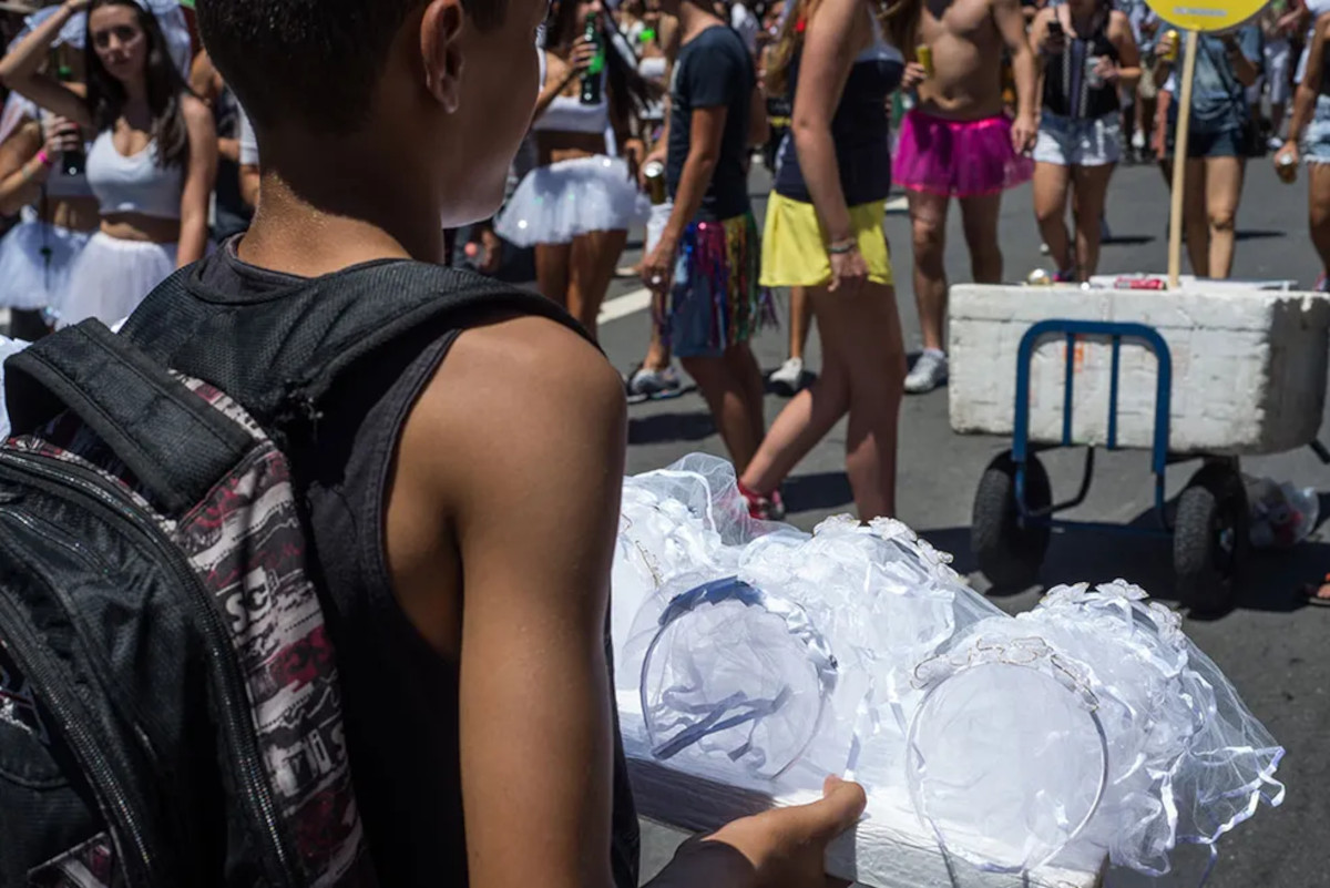 Imagem mostra um menino negro de costas vendendo adereços de carnaval na rua.