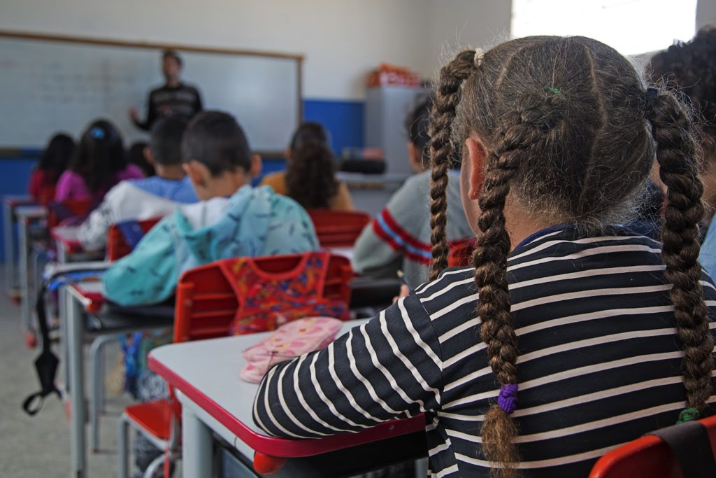 Imagem mostra uma menina negra e de tranças no cabelo de costas, sentada na sala de aula.