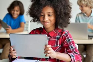 Imagem mostra uma menina negra sentada em sala de aula e usando um tablet. Atrás dela a dois meninos.