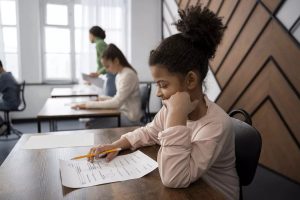 Imagem mostra uma menina negra segurando um lápis e olhando para uma folha a 4 na sala de aula.