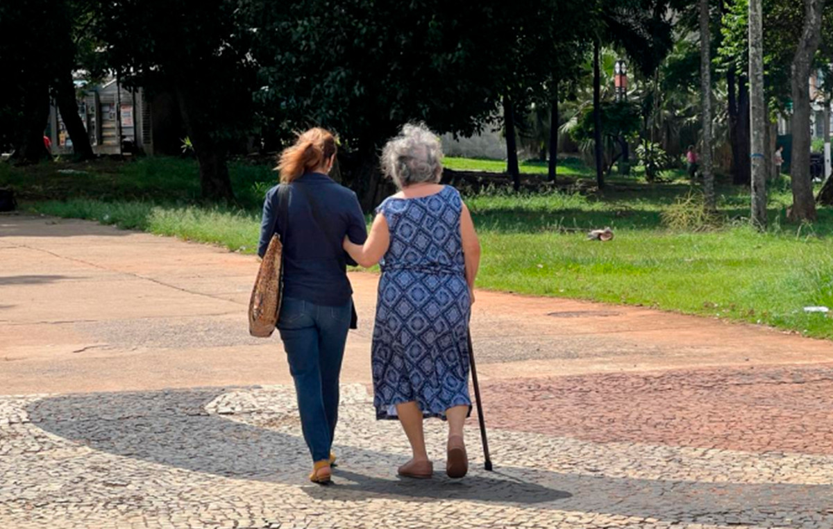 Imagem mostra uma mulher andando com uma idosa na rua. Elas estão de costas.