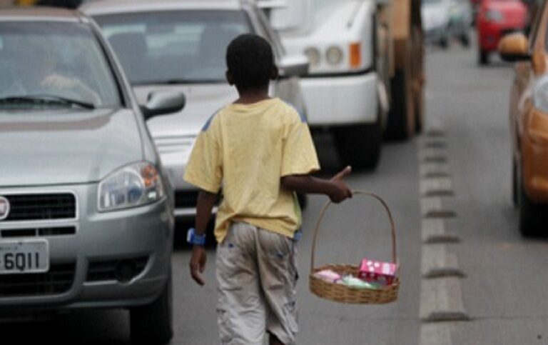 Imagem mostra um menino negro segurando uma cesta com doces para vender. Ele anda em meio a carros no trânsito.